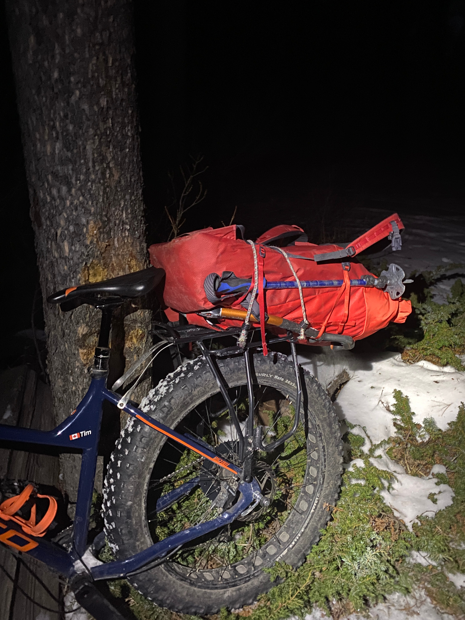 Red climbing pack and ice tools strapped directly to a fat-bike rack beside a tree at night.