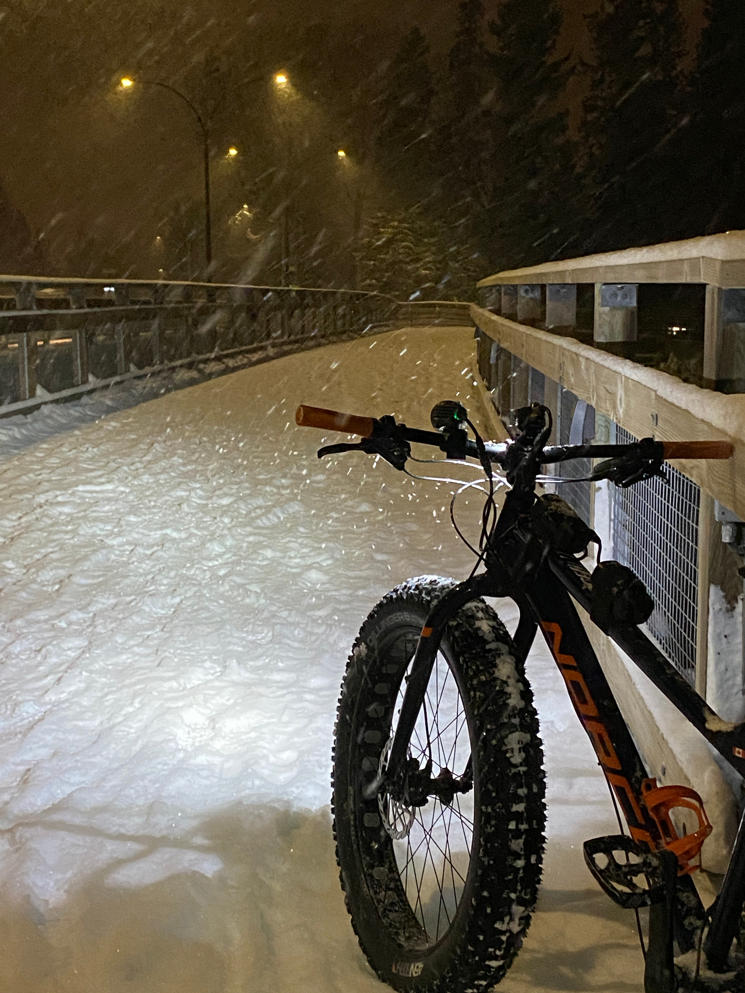 Close view of a fat bike on a snow-covered wooden bridge beneath streetlights at night.
