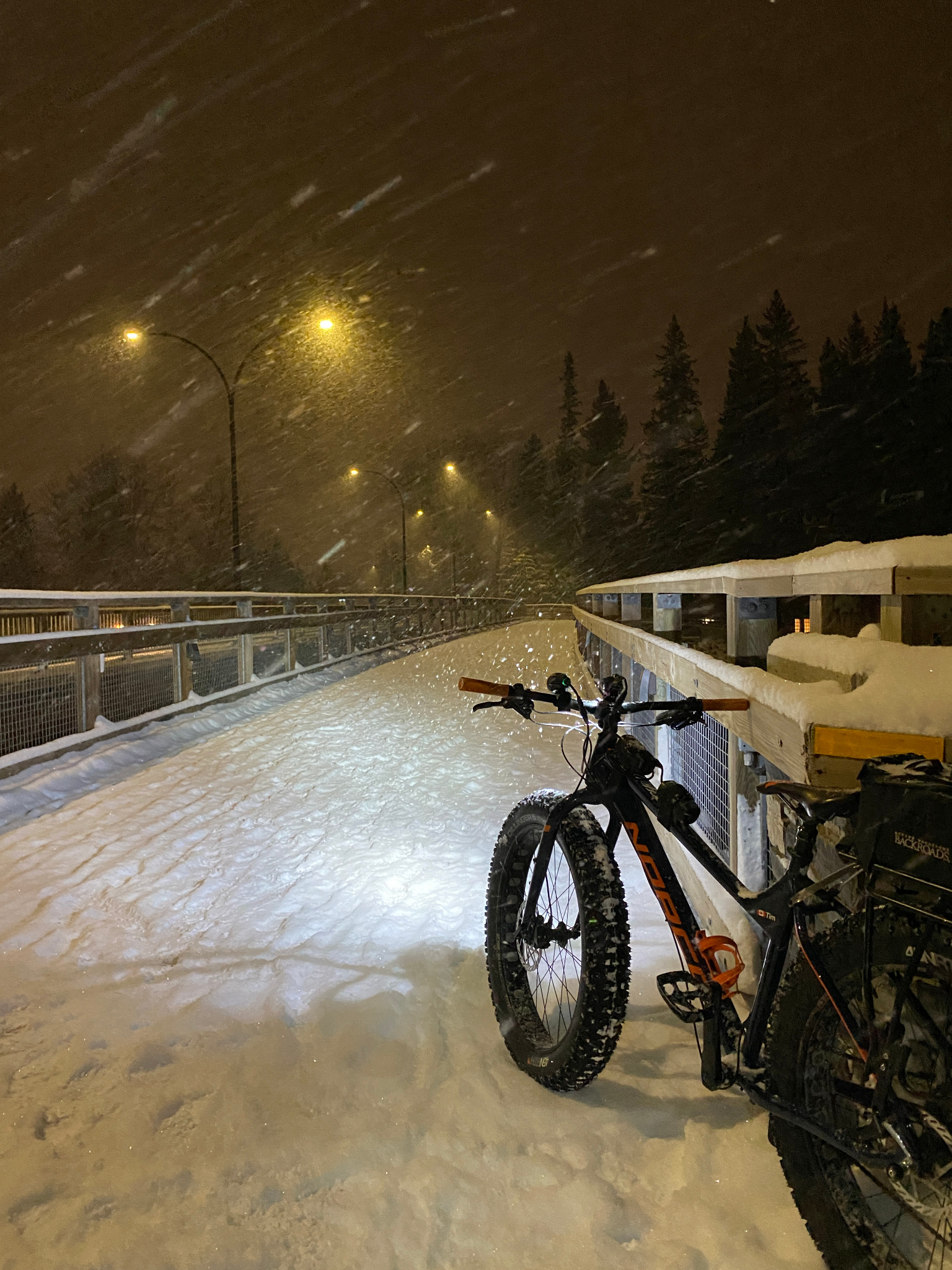 Loaded fat bike beside the railing of a snow-covered bridge during snowfall at night.