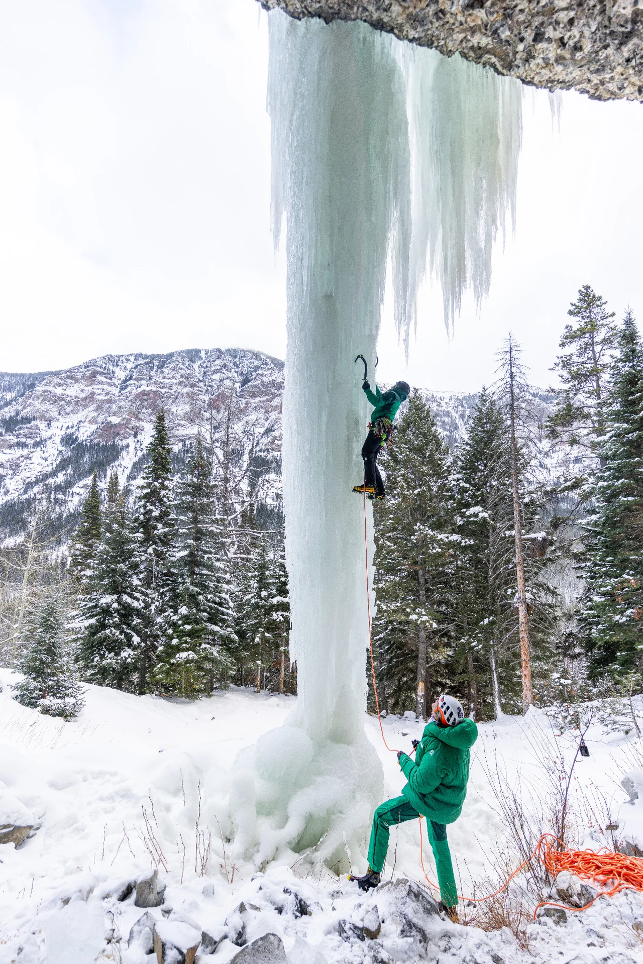 Andres Marin climbing Bingo Pillar above a belayer, with snowy trees behind the ice.