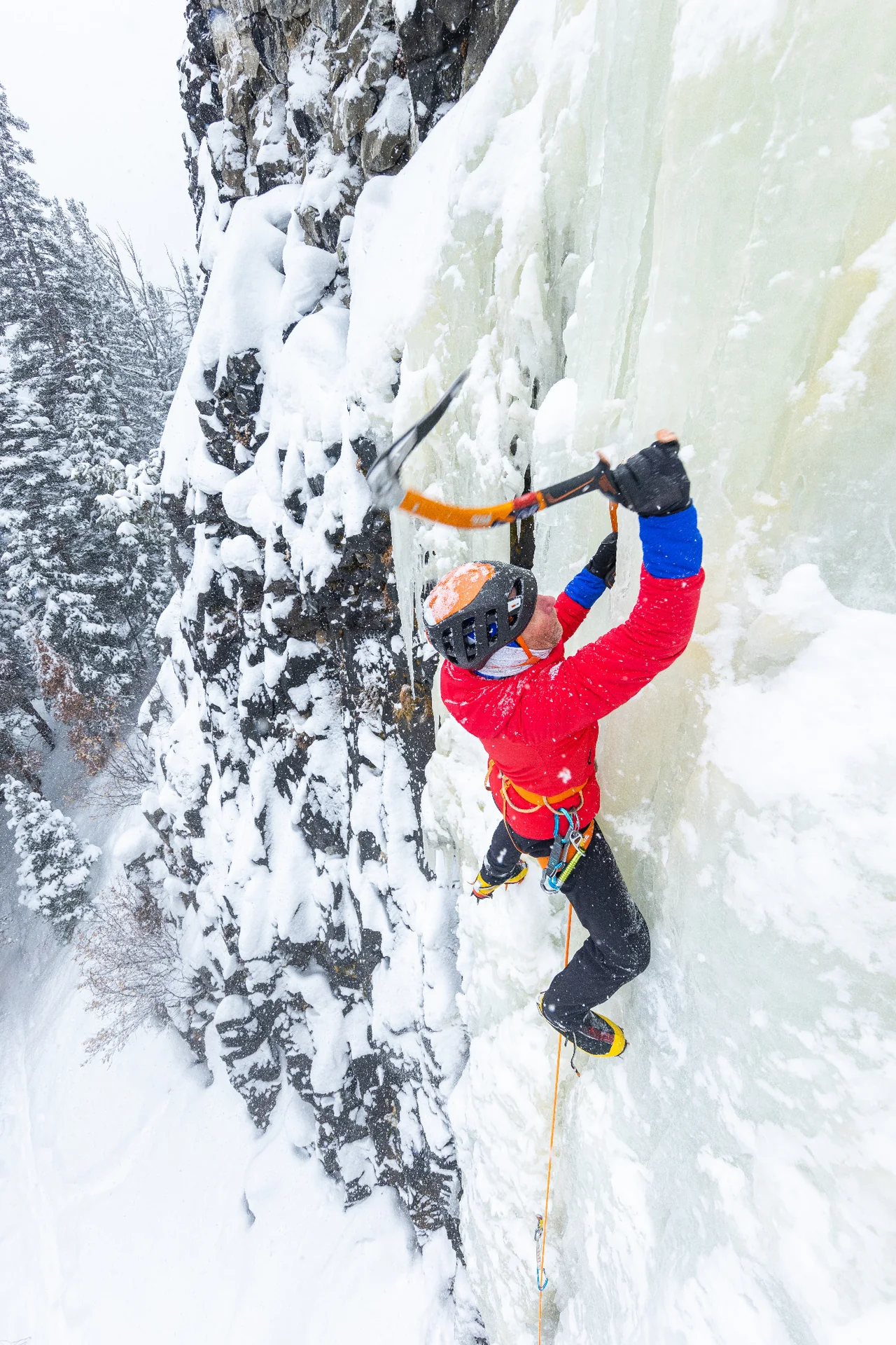 Conrad Anker raising an ice tool on The Good Looking One above snow-covered trees.