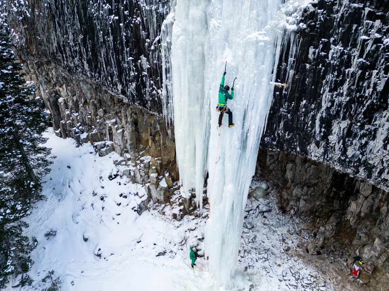 A climber in a green jacket ascends a broad ice pillar framed by dark rock walls and snow.