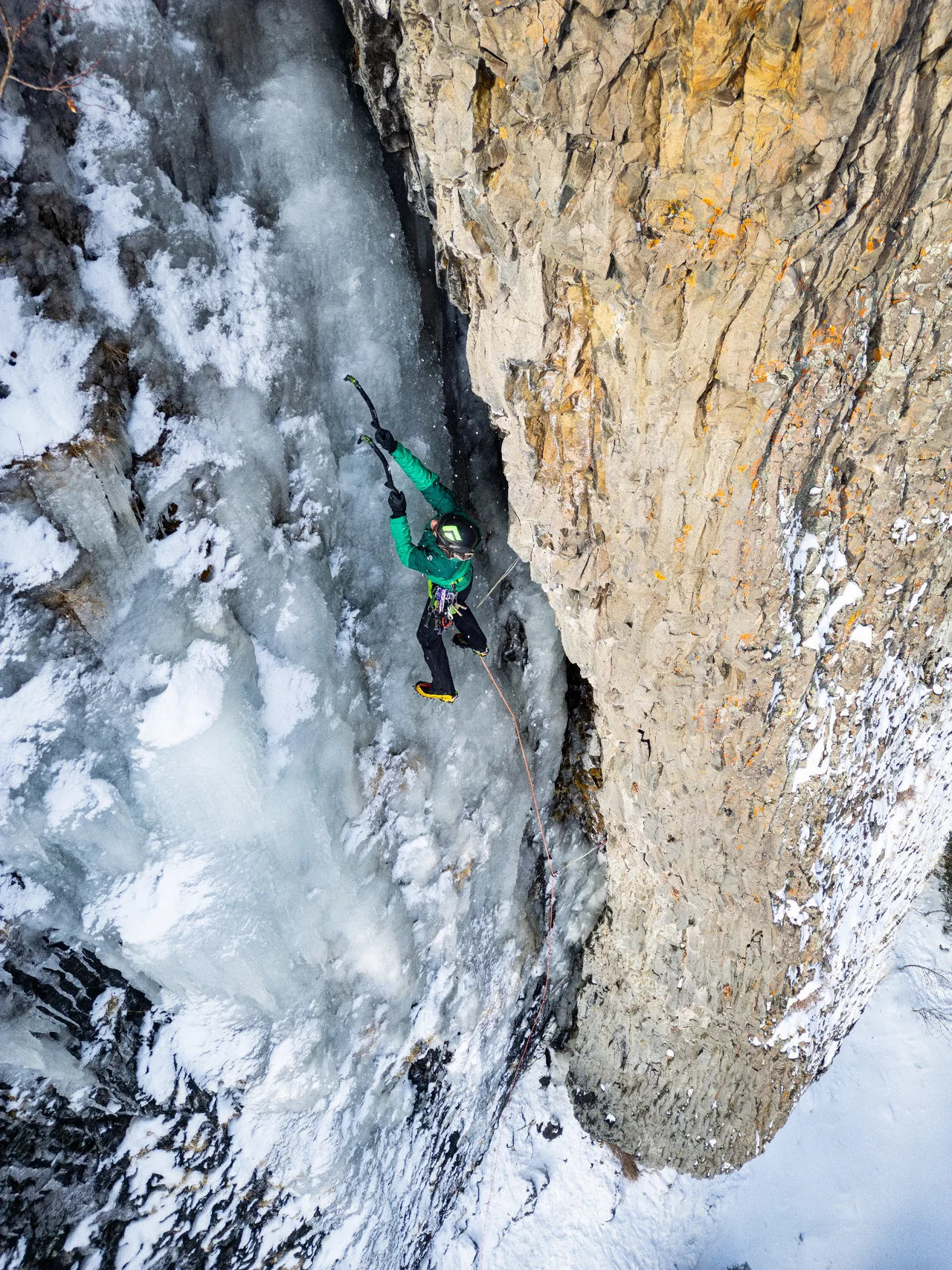 A climber in green climbs ice in a narrow corner beside a golden rock wall.