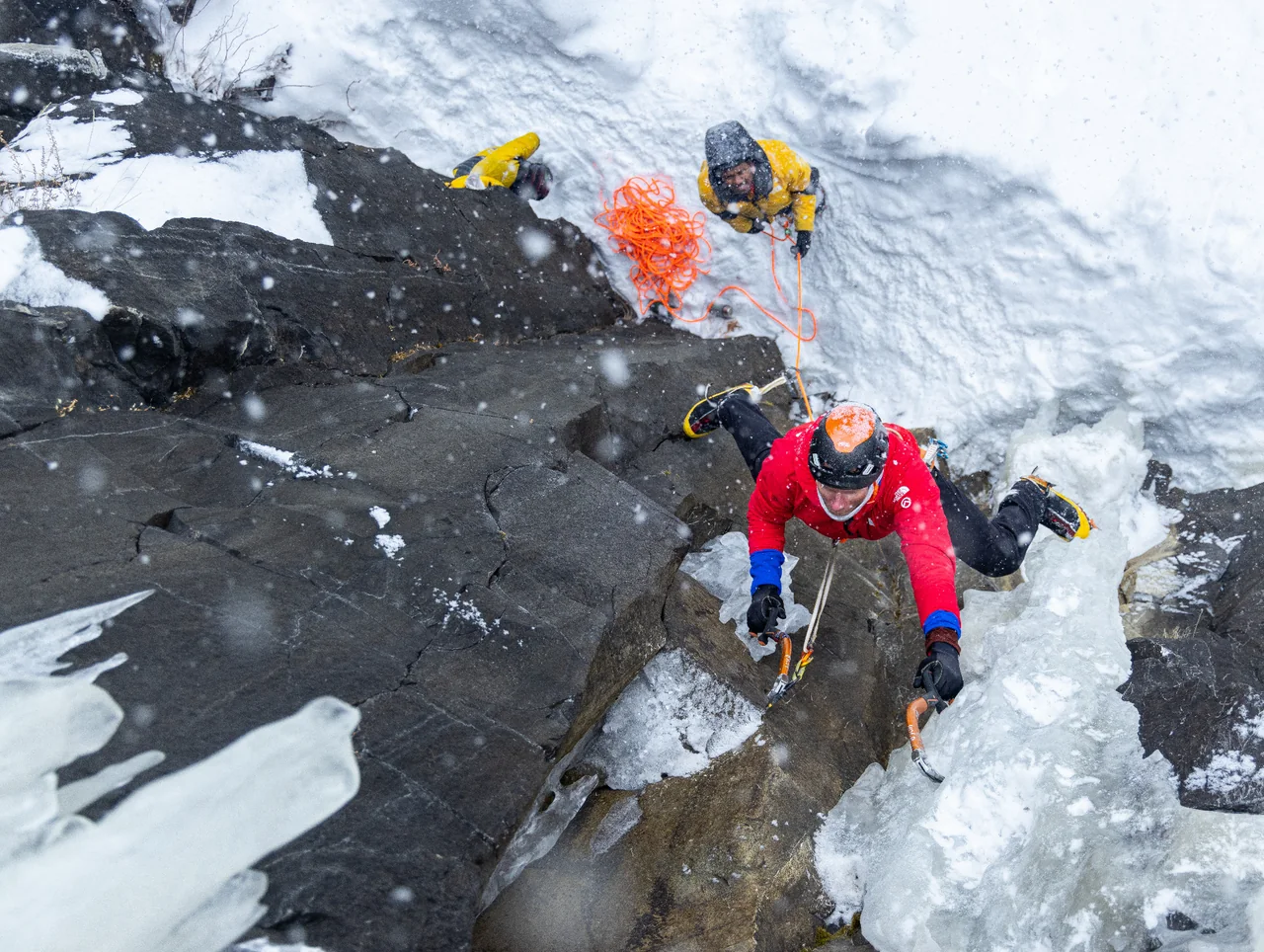A climber in red moves through a snowy rock-and-ice corner, with a belayer below.