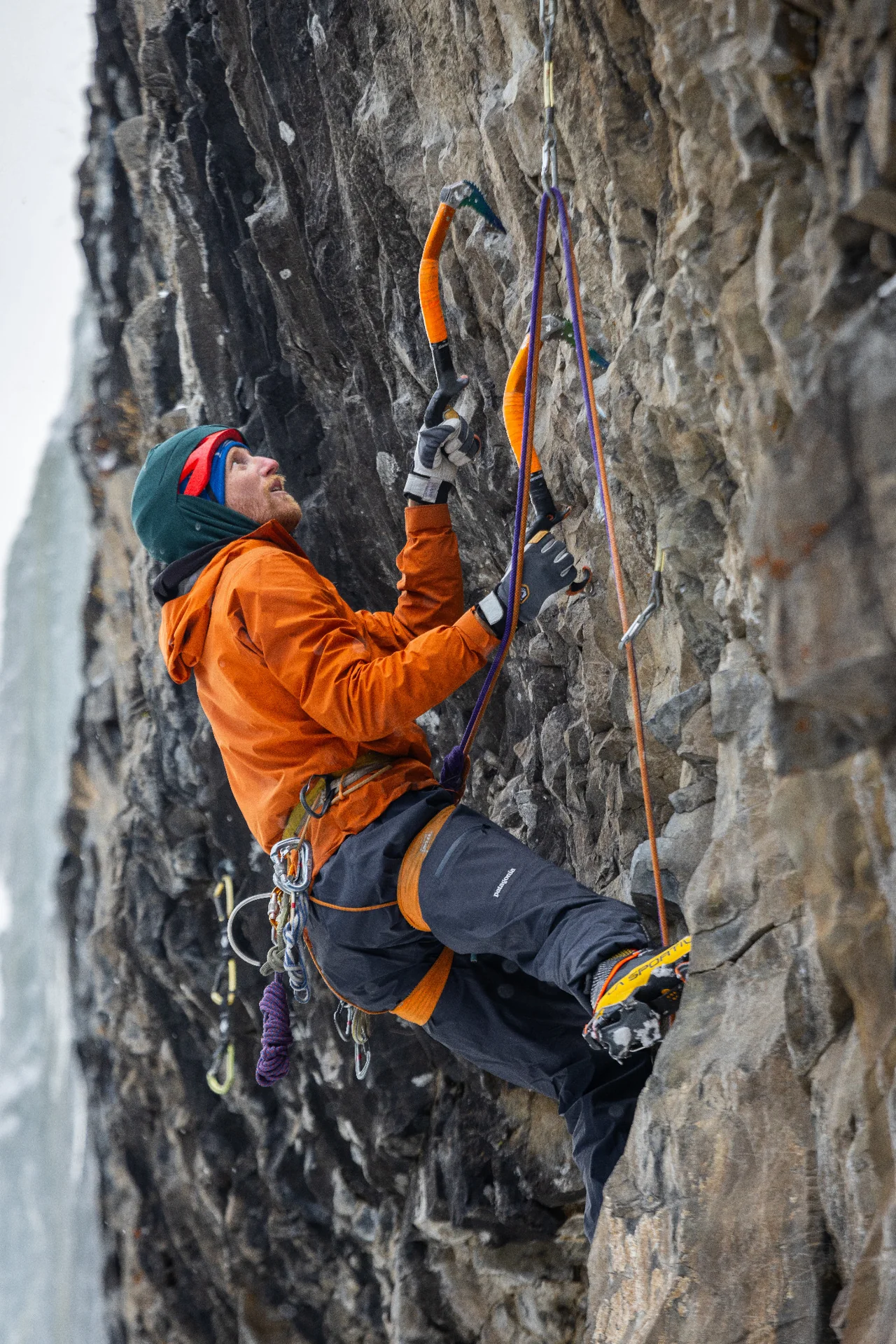 A climber in orange places ice tools on a steep rock wall.